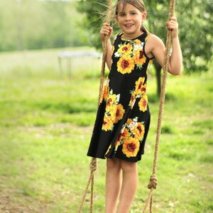 May include: A young person stands on a wooden swing, holding onto the rope supports. They are wearing a black dress adorned with large yellow sunflower patterns. The swing is suspended from a tree branch, set against a blurred green background.
