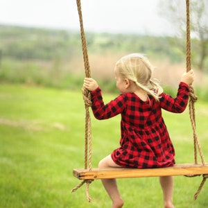May include: A young child sits on a wooden swing, facing away from the camera. The child wears a red and black plaid dress. The swing is suspended by thick rope from an unseen structure, set against a backdrop of green grass and a blurred landscape.