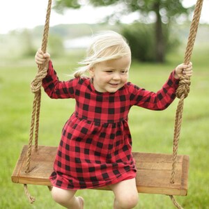 May include: A young child wearing a red and black plaid dress sits on a wooden swing with rope supports. The swing is suspended from a tree, and the background is a blurred green field.