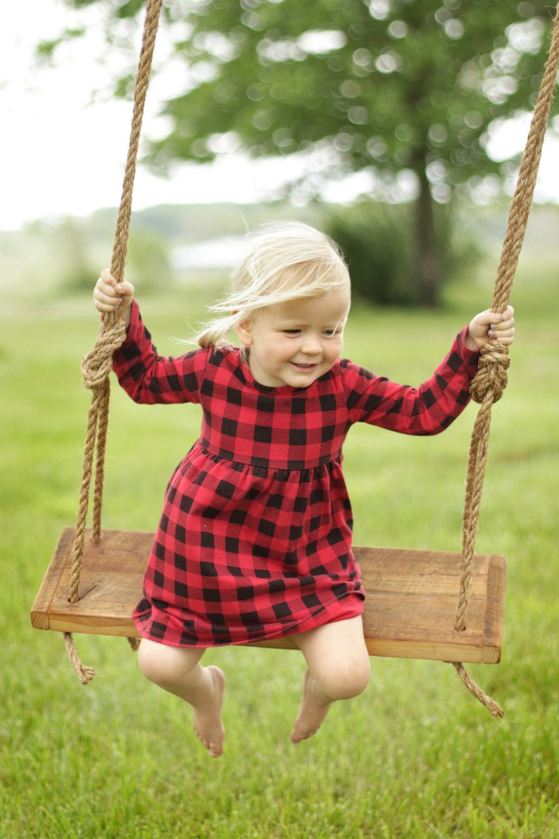 May include: A young child wearing a red and black plaid dress sits on a wooden swing with rope supports. The swing is suspended from a tree, and the background is a blurred green field.