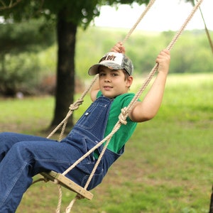May include: A young person is swinging on a wooden swing with rope supports. They are wearing a green shirt, blue denim overalls, and a camouflage baseball cap with the word "BOY" on it. The swing is hanging from a tree branch.