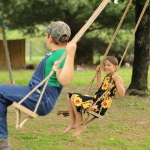 May include: Two children on wooden swings with rope supports. One child wears a green shirt and denim overalls, and a camouflage cap. The other child wears a black dress with a yellow sunflower pattern. The swings are in a grassy area.