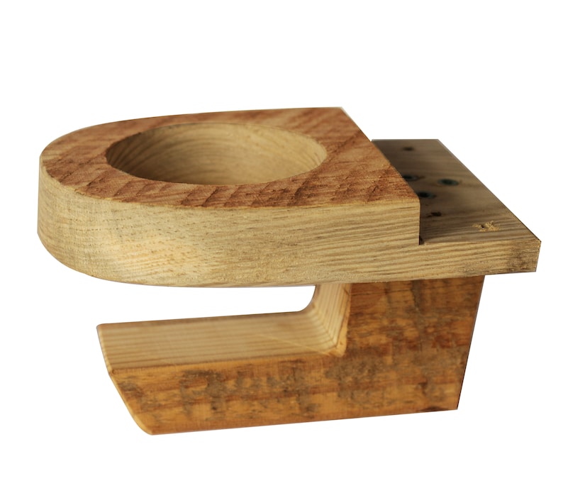 May include: A handcrafted wooden cup holder with a natural finish. The holder features a circular indentation for a cup and a rectangular base. The wood grain is visible, adding to its rustic charm. The holder is designed to be mounted on a surface.