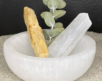 May include: A white selenite bowl with a palo santo stick and a selenite wand inside. A sprig of eucalyptus is also in the bowl.