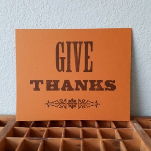 May include: Orange cardstock sign with the text "GIVE THANKS" in brown lettering with a decorative flourish below the text.
