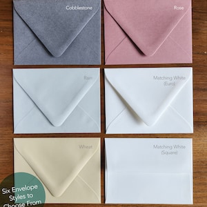 Puede incluir: Seis estilos diferentes de sobres mostrados en una superficie de madera. Los colores son Cobblestone (gris), Rain (azul claro), Rose (rosa), Wheat (crema), Matching White (Euro) y Matching White (Square).