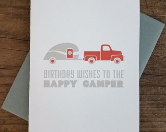 May include: A white card with a red truck pulling a gray camper. The text on the card reads "Birthday Wishes to the Happy Camper".