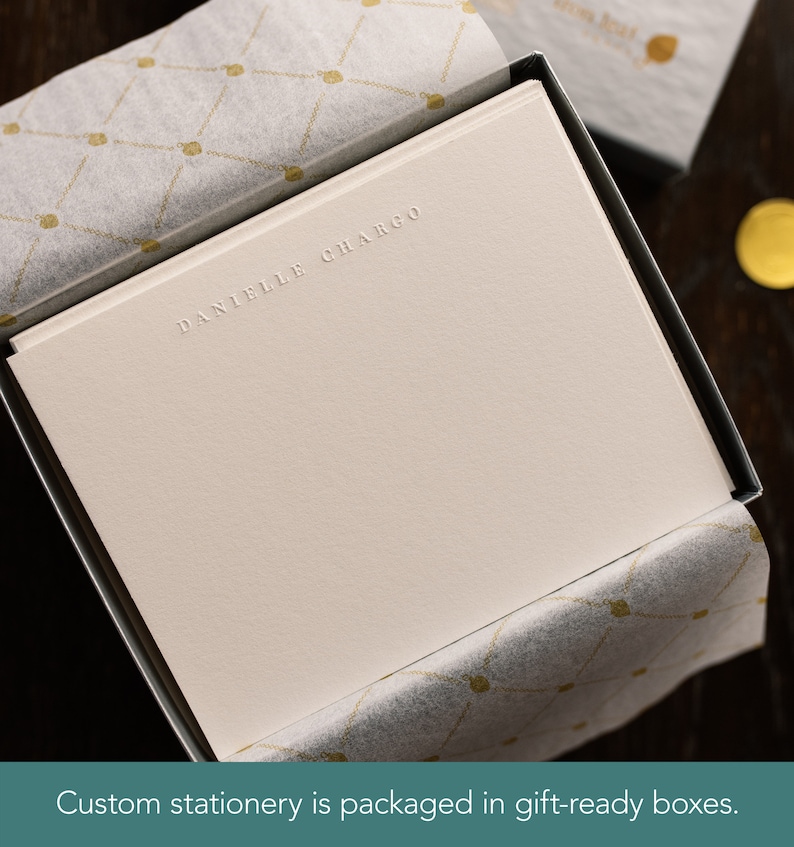 Puede incluir: Una caja blanca con una tarjeta blanca en su interior. La tarjeta tiene el nombre "DANIELLE CHARGO" en relieve en letras doradas. La caja est&aacute; forrada con papel de seda blanco con un patr&oacute;n dorado.
