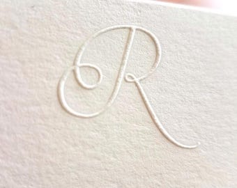 May include: A cream-colored cardstock paper featuring a debossed, elegant cursive letter R. The texture of the paper is visible, showing a fine, fibrous grain. The letter is pressed into the surface, creating a subtle, recessed effect.
