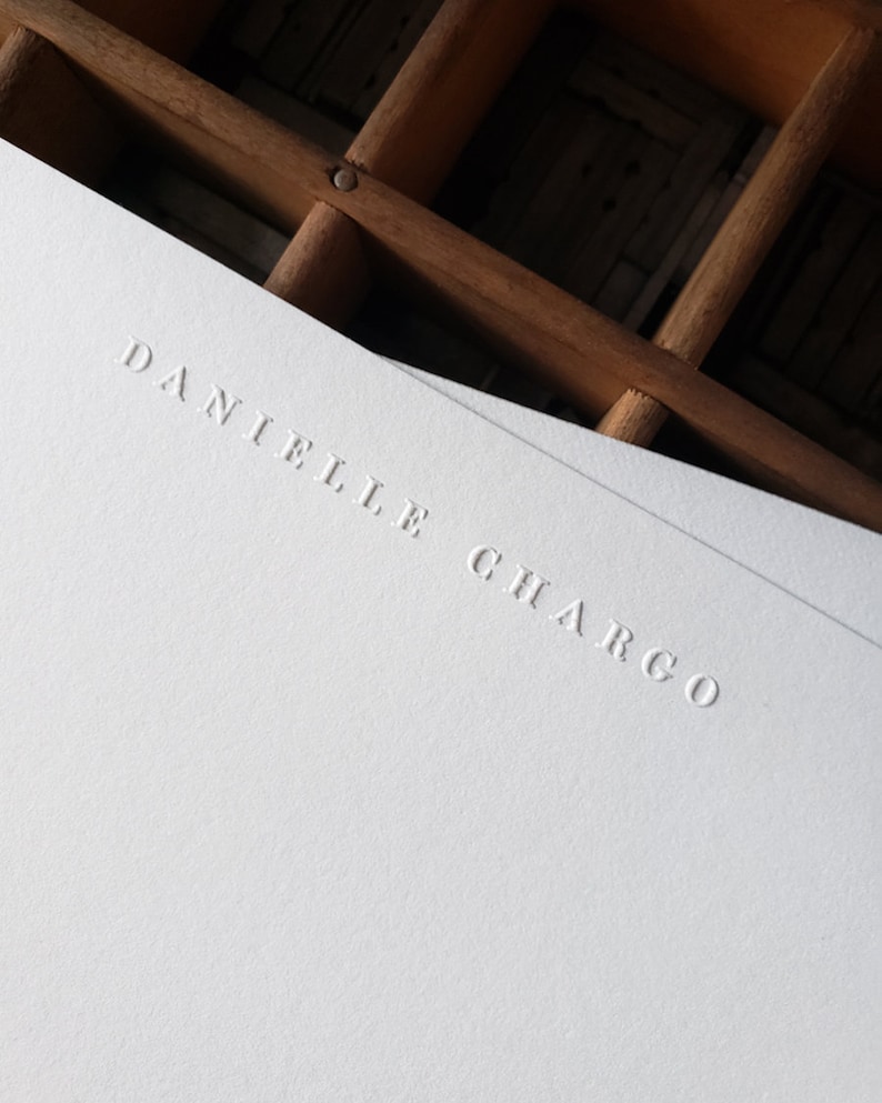 Puede incluir: Cartulina blanca con el nombre "Danielle Chargo" en relieve en una fuente simple y elegante.