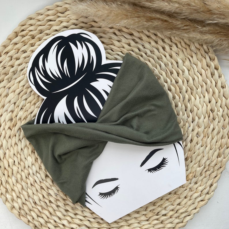 May include: A green fabric headband with a twist design. The headband is on a white background with a black and white illustration of a woman with her hair in a bun.