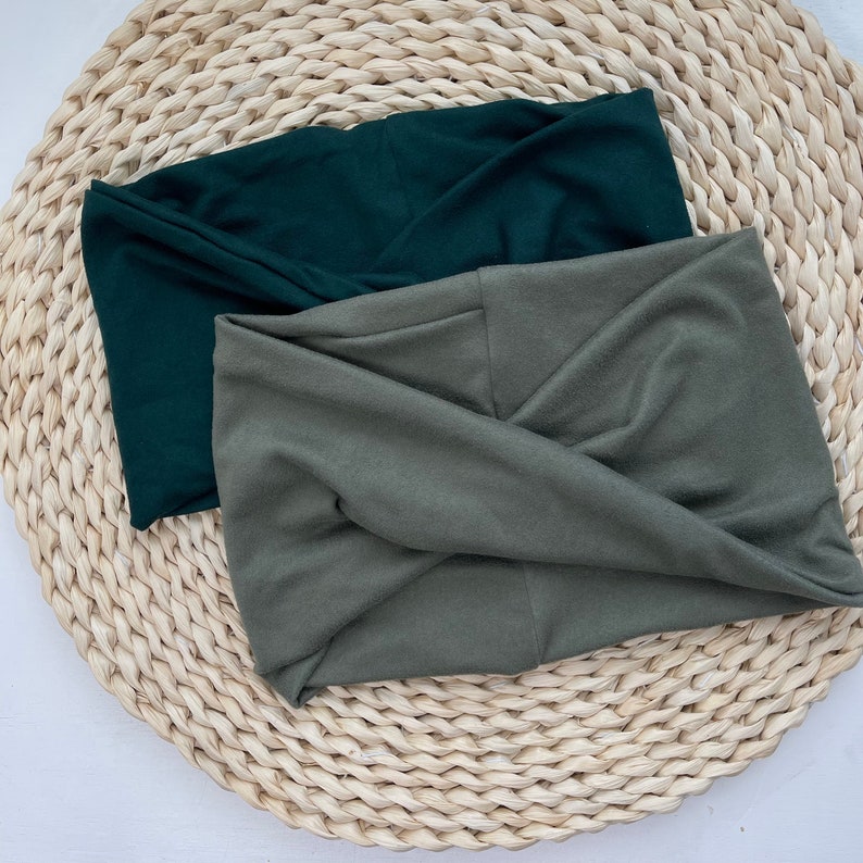 May include: Two fabric headbands, one dark green and one olive green, are folded and displayed on a woven straw surface.