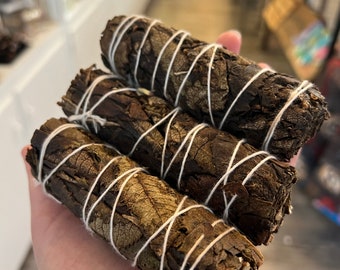 May include: Four bundles of dried leaves, tightly wrapped with white string. The leaves are a dark brown color, with visible veins and textures. The bundles are held in a hand, suggesting a natural product.