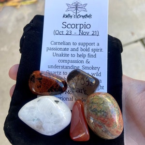 May include: A black velvet pouch containing a set of crystals for the zodiac sign Scorpio. The crystals are carnelian, unakite, smoky quartz, and a green and red stone. The pouch has a tag that reads "Kelly's Crystals Scorpio (Oct 23 - Nov 21)".