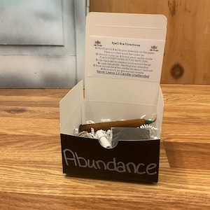 May include: An open white box with a black front panel, labeled "Abundance" in white script. Inside are various items, including a brown stick, a small black square, and a rolled paper. The box also contains instructions.
