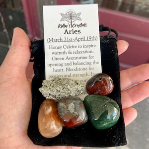 May include: A black velvet pouch containing a set of crystals for the zodiac sign Aries. The crystals are honey calcite, green aventurine, bloodstone, and pyrite. The pouch has a tag that reads "Kelly's Crystals Aries (March 21st-April 19th)" and describes the properties of each crystal.