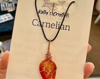 May include: A carnelian pendant necklace with a copper wire wrap. The pendant is a natural, orange-brown stone. The necklace is on a black cord and has a dragonfly charm. The card reads "Kelly's Crystals, Carnelian, The Motivator".