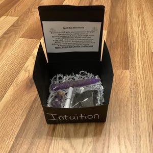 May include: A black box with the word "Intuition" written on the front. The box is open and contains a purple candle, a small jar with crystals, a white pen, and a small bag of black herbs. The box also includes a piece of paper with instructions for a spell box.