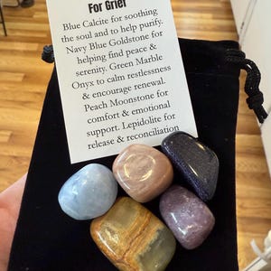 May include: A black velvet pouch containing six polished gemstones. A card reads "Kelly's Crystals For Grief" and lists the properties of each stone: Blue Calcite, Navy Blue Goldstone, Green Marble Onyx, Peach Moonstone, and Lepidolite.