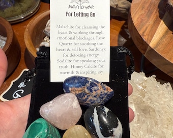 May include: A black velvet pouch holding five polished gemstones: malachite, rose quartz, sardonyx, sodalite, and honey calcite. A small card reads "Kelly's Crystals For Letting Go", with descriptions of each stone's properties.