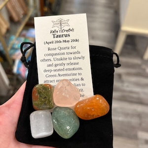 May include: A black velvet pouch containing five tumbled crystals: green aventurine, rose quartz, unakite, clear quartz, and carnelian. A card with the text "Kelly's Crystals Taurus (April 20th-May 20th) Rose Quartz for compassion towards others. Unakite to slowly and gently release deep-seated emotions. Green Aventurine to attract opportunities &" is visible.