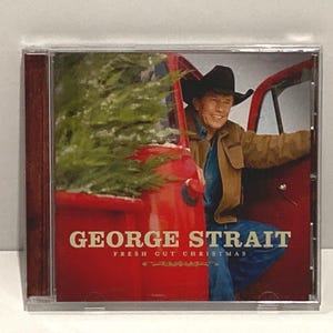 May include: A 'George Strait Fresh Cut Christmas' CD with A red pickup truck with a green tree in the bed.  George Strait is wearing a brown jacket and a cowboy hat is leaning out of the truck.