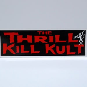 May include: A rectangular sticker with the text "THE THRILL KILL KULT" in red, bold letters on a black background. A white silhouette of a devil figure with a trident is on the right side of the sticker. The sticker has a white border.