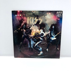 May include: Album cover for the band KISS, titled "Alive!" The cover features the band members in full stage makeup and costumes, playing their instruments on a stage with a dark background. The band's name is in large, illuminated letters.