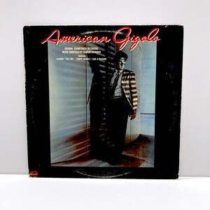 May include: Vintage vinyl record album cover for "American Gigolo." The cover features the title in red script, with the original soundtrack recording information. The image shows a man in a suit, with the names of the featured artists and songs.