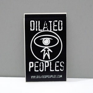 May include: Black rectangular sticker with white text and a circular graphic. The text reads "DILATED PEOPLES" above and below a white eye-shaped symbol. The website address www.DILATEDPEOPLES.com is at the bottom.