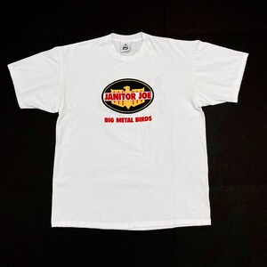 May include: White t-shirt with a crew neck featuring a graphic design. The design includes the text "JANITOR JOE" in red and white, set within a black oval, with the words "BIG METAL BIRDS" in red below.
