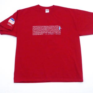 May include: A red t-shirt with the text "HEARTSTOPPINPANTSDROPPING...LEGENDARY...E STREET BAND" in white and light blue. The left sleeve has a circular patch with the word "ADLANDS" in a white and blue design.