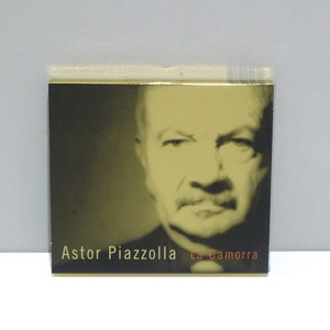 May include: Square CD case featuring a sepia-toned portrait of Astor Piazzolla. The text "Astor Piazzolla" is above "La Camorra" in red. The case has a gold border and is set against a light gray background.