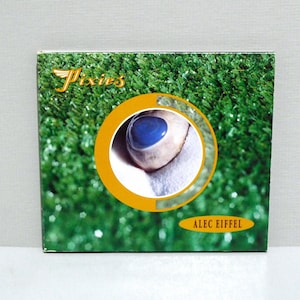 May include: Square album cover featuring a close-up of an eye with a bright blue iris, set against a white background within a yellow circle. The background is green, resembling grass. The word "Pixies" is in gold lettering, and "ALEC EIFFEL" is in a yellow rectangle.