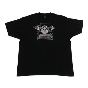 May include: Black t-shirt featuring a white line drawing of a vintage typewriter. The typewriter illustration is centered on the shirt and includes details of the keys, carriage return lever, and paper feed. The shirt is made of a soft material.