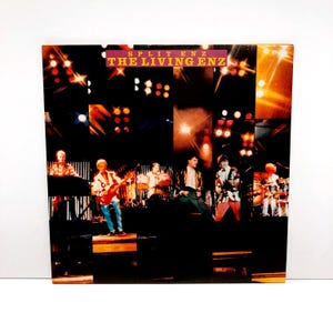 May include: Split Enz album cover, "The Living Enz", featuring a band performing on stage. The album cover has a collage of images with the band members playing instruments. The album title is in a maroon banner at the top.