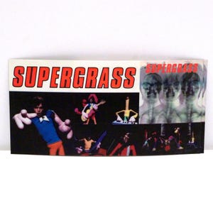 May include: A Supergrass album cover featuring the band's name in red block letters. The cover art includes cartoon-like figures in various poses, with a black background and a white border. The right side of the cover has a blurred image of three faces.
