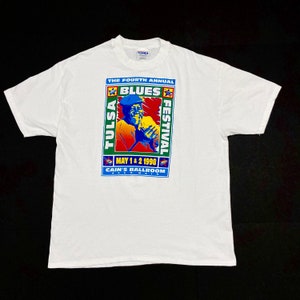 May include: White t-shirt featuring a colorful graphic for the Fourth Annual Tulsa Blues Festival, held May 1 & 2, 1998, at Cain's Ballroom. The design includes a portrait of a musician and the event details.