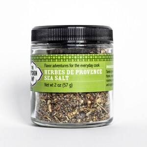 May include: A glass jar with a black lid filled with a blend of herbs and sea salt. The label reads "The Kitchen  Imp" and "Herbes de Provence Sea Salt" with a net weight of 2 ounces (57 grams).