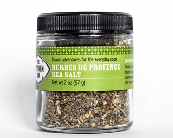 Può includere: Un barattolo di vetro con un coperchio nero riempito di un mix di erbe e sale marino. L'etichetta riporta "The Kitchen Imp" e "Herbes de Provence Sea Salt" con un peso netto di 2 once (57 grammi).