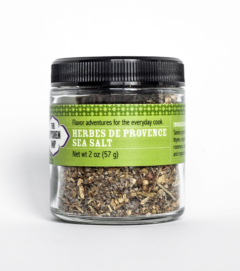 May include: A glass jar with a black lid filled with a blend of herbs and sea salt. The label reads "The Kitchen  Imp" and "Herbes de Provence Sea Salt" with a net weight of 2 ounces (57 grams).
