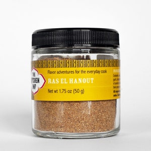 May include: A glass jar with a black lid filled with Ras El Hanout spice blend. The label is yellow with black text and reads "The Kitchen Imp" and "Flavor adventures for the everyday cook". The jar contains 1.75 ounces (50 grams) of spice.