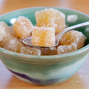 May include: A teal and green ceramic bowl filled with sugared ginger cubes. A silver spoon holds a piece of the candied ginger. The ginger is a light amber color and coated in white sugar crystals. The bowl sits on a light brown surface.