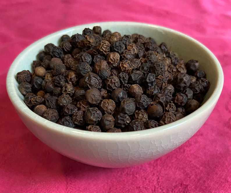 May include: A white ceramic bowl filled with black peppercorns. The peppercorns are round and have a slightly wrinkled texture.