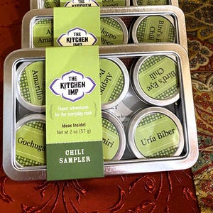 May include: A chili sampler set in a metal tin, with small round tins featuring green labels. The labels display various chili names. A green card with "The Kitchen Imp" logo and "Chili Sampler" text is in front.