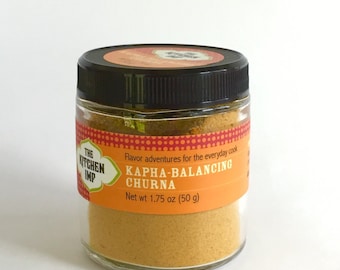 May include: A glass jar with a black lid containing a yellow powder. The label reads "The Kitchen Imp" and "Kapha-Balancing Churna". The label also states "Flavor adventures for the everyday cook" and "Net wt 1.75 oz (50 g)".