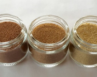 Könnte beinhalten: Drei Glasbehälter gefüllt mit Sand in verschiedenen Farben. Die Behälter sind in einer Reihe von links nach rechts angeordnet. Der Sand im linken Behälter ist braun, der Sand im mittleren Behälter ist hellbraun und der Sand im rechten Behälter ist gelb.