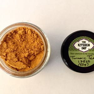 Peut inclure: Un pot en verre rempli d'une poudre brune étiquetée "Turmeric Tea" par "The Kitchen Imp". L'étiquette indique également "Flavor adventures for the everyday cook" et "1.75 oz (50g)".
