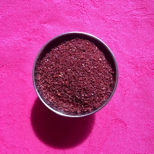 May include: A small metal container filled with a deep red, finely ground spice. The container is sitting on a pink fabric surface.