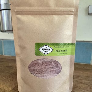 May include: A brown paper bag of Kala Namak salt. The label reads "The Kitchen Imp" and "Flavor adventures for every cook." The bag has a clear oval window showing the pinkish salt. It contains 10 oz (283 g) of salt.